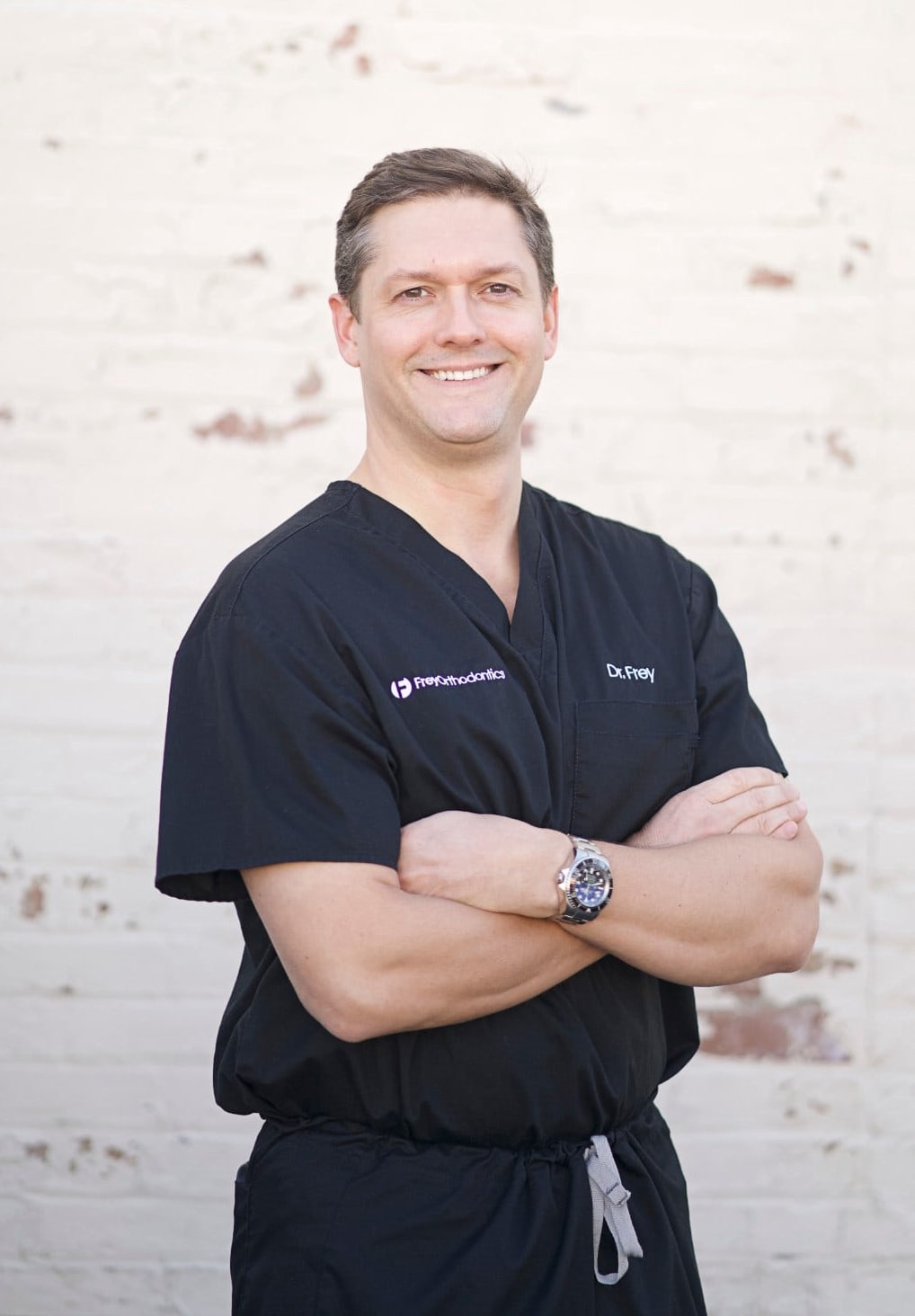 Dr. Dwight Frey, orthodontist in Naperville, wearing black scrubs with Frey Orthodontics logo, smiling confidently against a light brick background.